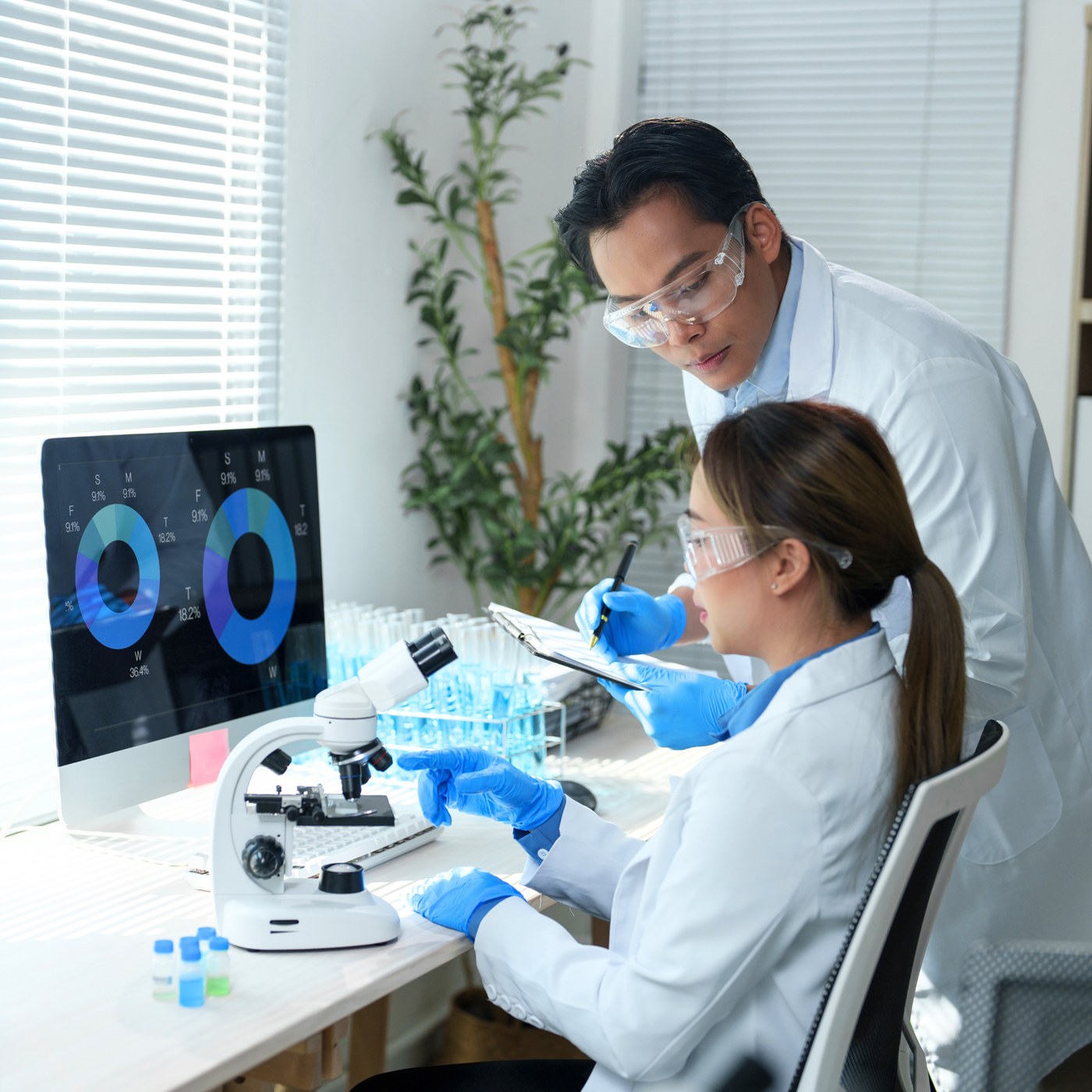 Two research scientists in front of a computer and microscope with vials beside it